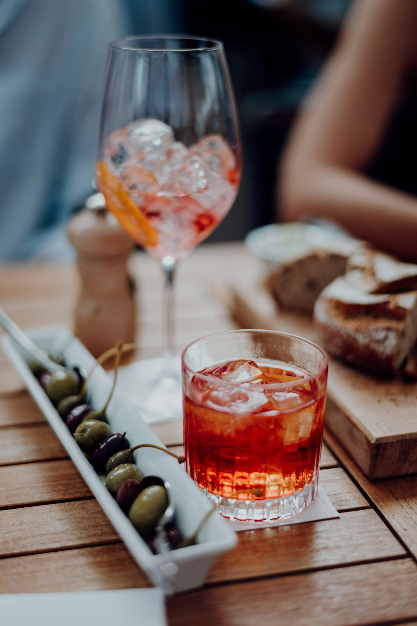 Czerwony aperitif bezalkoholowy z lodem w szklance oraz kieliszek koktajlowy i oliwki podane na drewnianym stole.