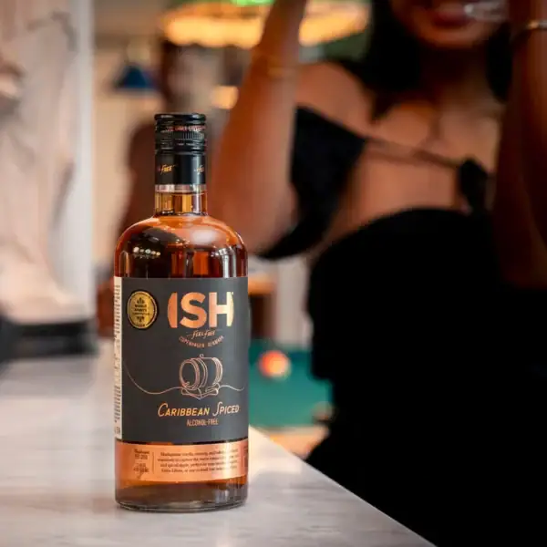 Butelka bezalkoholowej alternatywy rumu ISH Caribbean Spiced Spirit na marmurowym blacie w nastrojowym barze z rozmytym tłem ludzi.