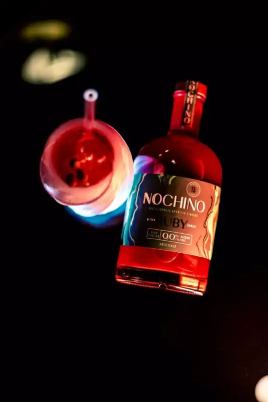 Butelka bezalkoholowego aperitifu Nochino Ruby i szklanka z czerwonym drinkiem ułożone na czarnym tle w intensywnym podświetleniu.