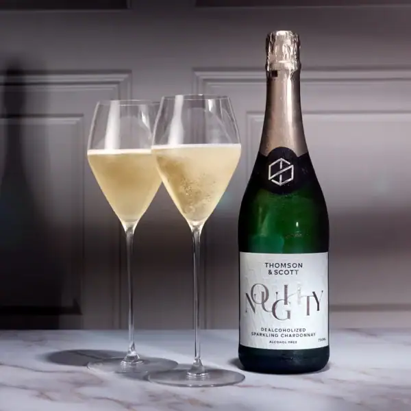 Butelka bezalkoholowego wina musującego Thomson i Scott Noughty Sparkling Chardonnay obok dwóch napełnionych kieliszków