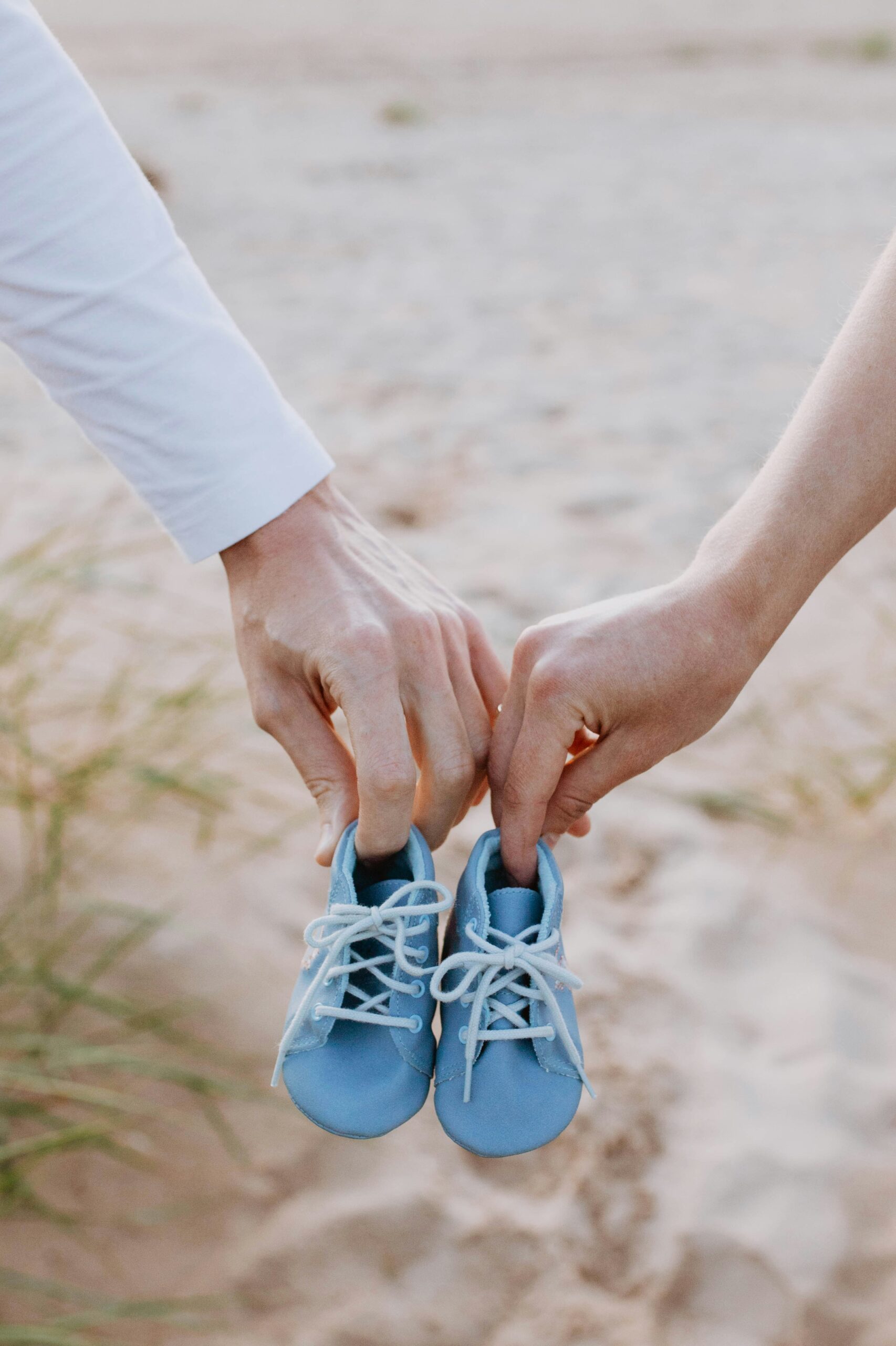 Dwie dłonie dorosłych osób trzymają wspólnie parę małych, niebieskich bucików dziecięcych na tle piaszczystej plaży. Bezpieczne wino bezalkoholowe na baby shower to wspaniały sposób na wzniesienie toastu za przyszłą mamę.