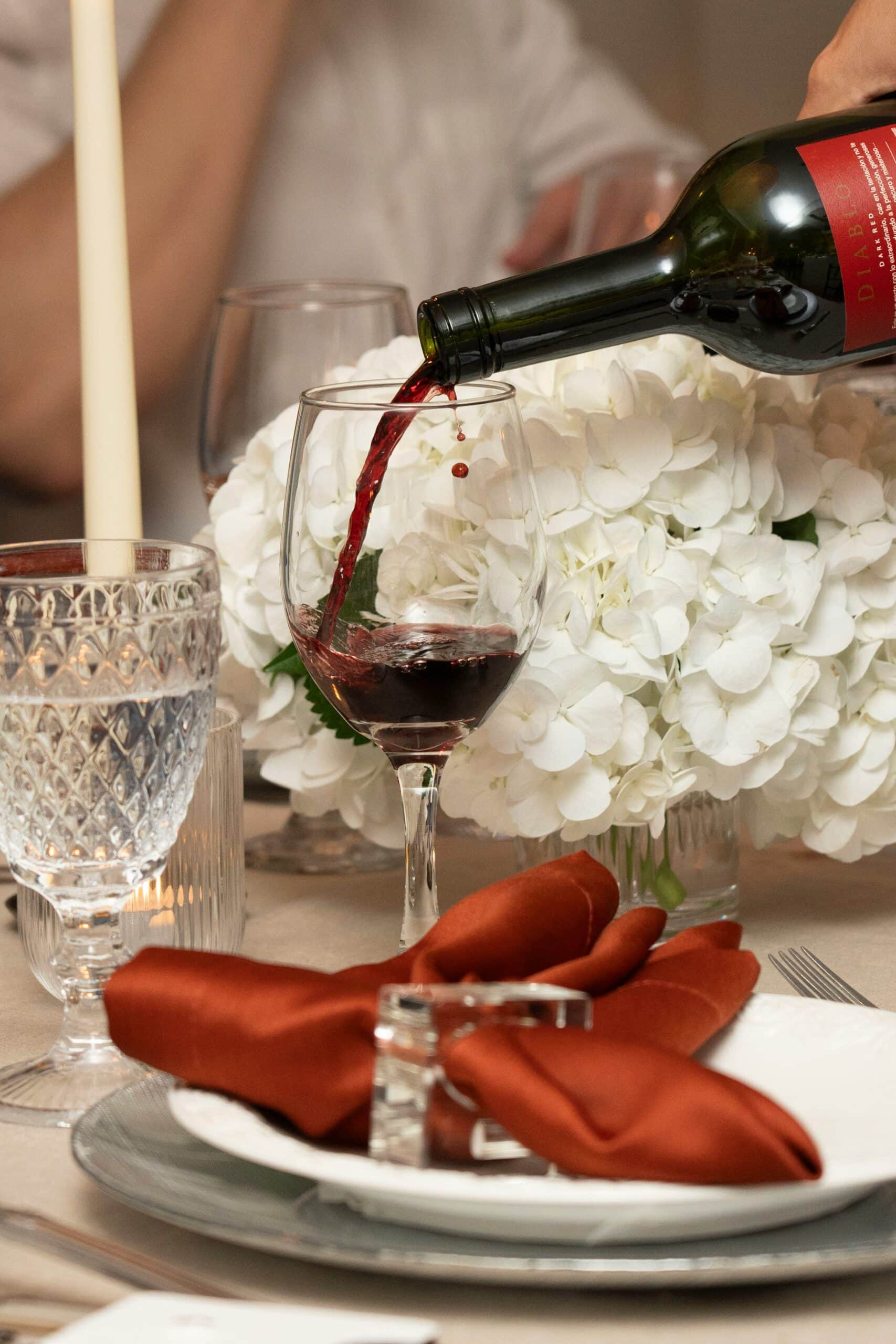 Czerwone wino bezalkoholowe na prezent nalewane z butelki do kieliszka na elegancko zastawionym stole z białymi kwiatami.